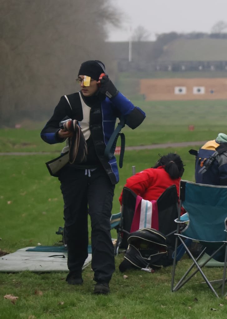 Photo of me walking across an outdoor shooting range wearing a shooting jacket and sling.